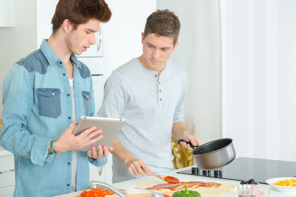Two young men in a kitchen, one following a recipe on a tablet while the other spreads sauce on pizza dough.